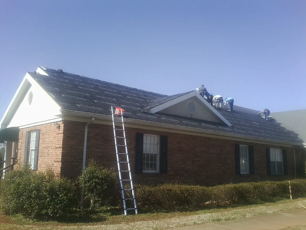 Crew tearing shingles off a brick ranch roof with a ladder set — Metal Roof Repair work by our crew in Champion
