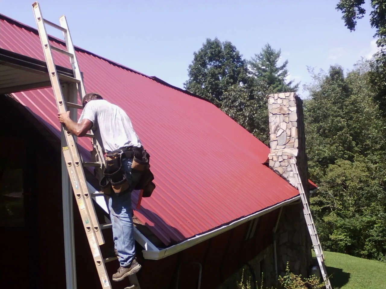 Roofer on a ladder beside a red metal roof and stone chimney — Soffit & Fascia Repair by Roofing Brothers in Champion